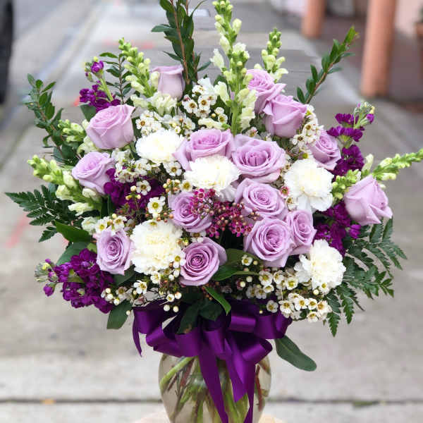 Lavender roses and white flowers arranged in a glass vase with a purple ribbon.