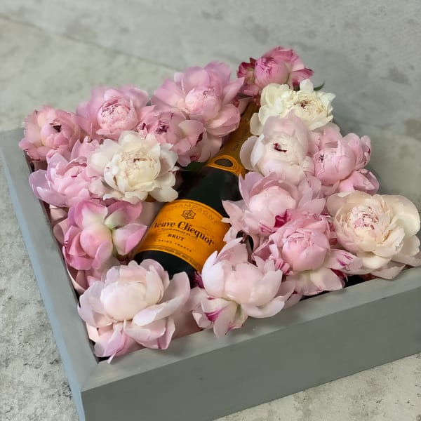 Pink and white peonies arranged around a champagne bottle in a gray box