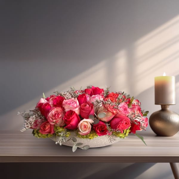 Low bowl arrangement of pink and red roses with a candle beside it