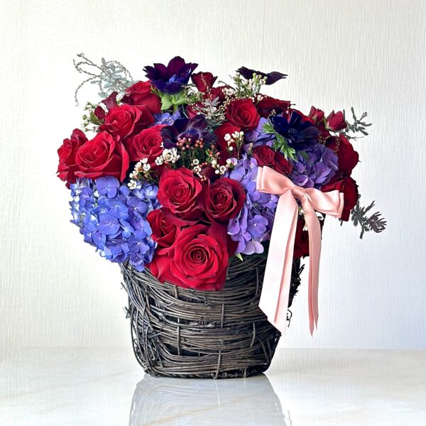 Bouquet of red roses and blue hydrangeas in a woven basket with a pink ribbon