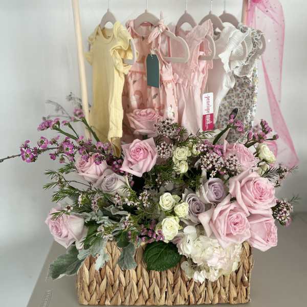 Pink and white rose arrangement in a woven basket with baby dresses behind it