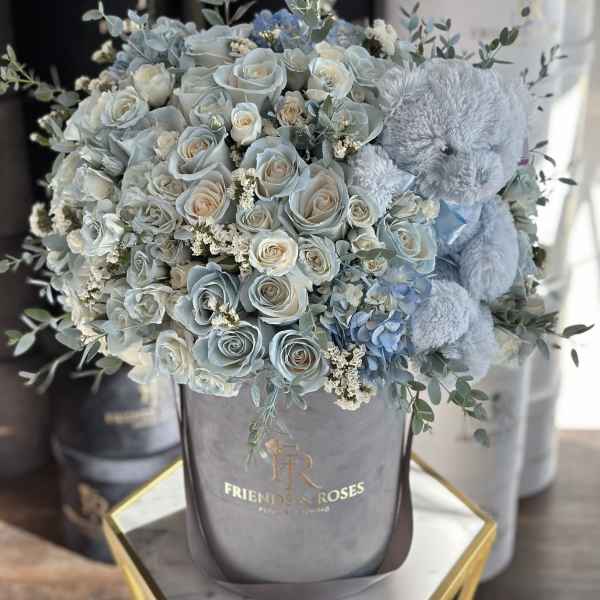 Blue roses and hydrangeas in a gray hatbox with a teddy bear