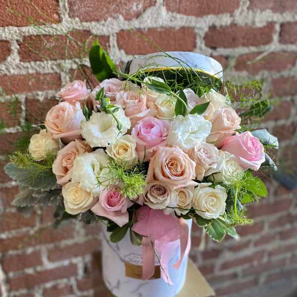 Round hatbox of blush and cream roses with white blooms and greenery against a brick wall