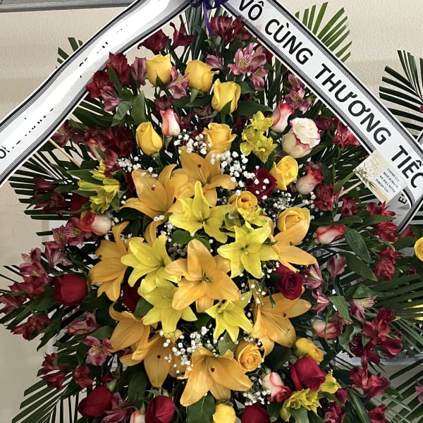 Large standing floral wreath with yellow lilies, red roses, and a purple ribbon