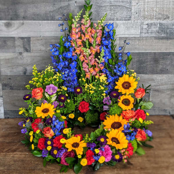 Tall circular wreath with bright sunflowers, roses, carnations, and blue delphinium.
