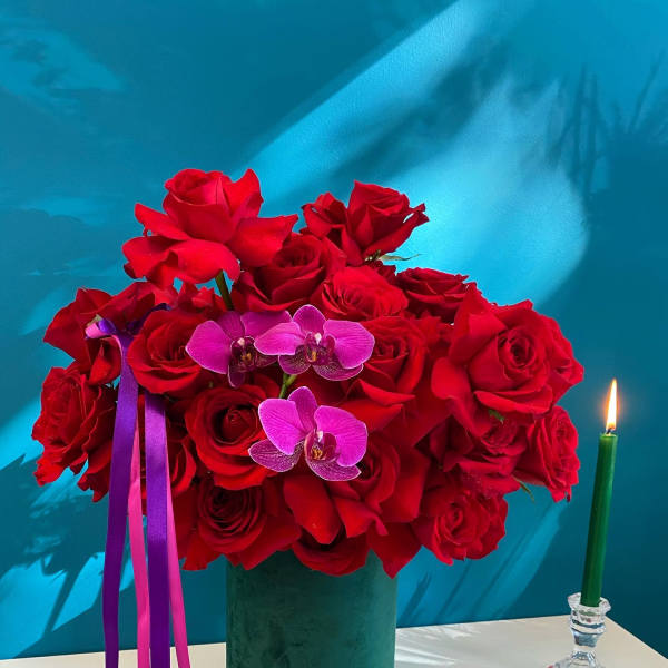 Red roses and purple orchids in a green vase with ribbon accents