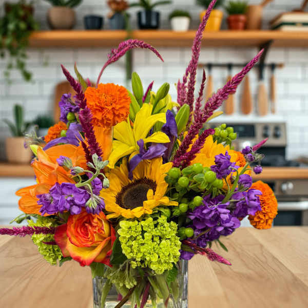 Bright mixed bouquet in a square glass vase with sunflower, roses, and purple blooms