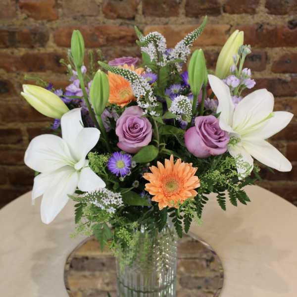 Mixed bouquet with white lilies, lavender roses, and orange gerbera daisies in a glass vase