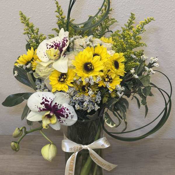 Yellow sunflowers and white orchids in a glass vase with a birthday ribbon