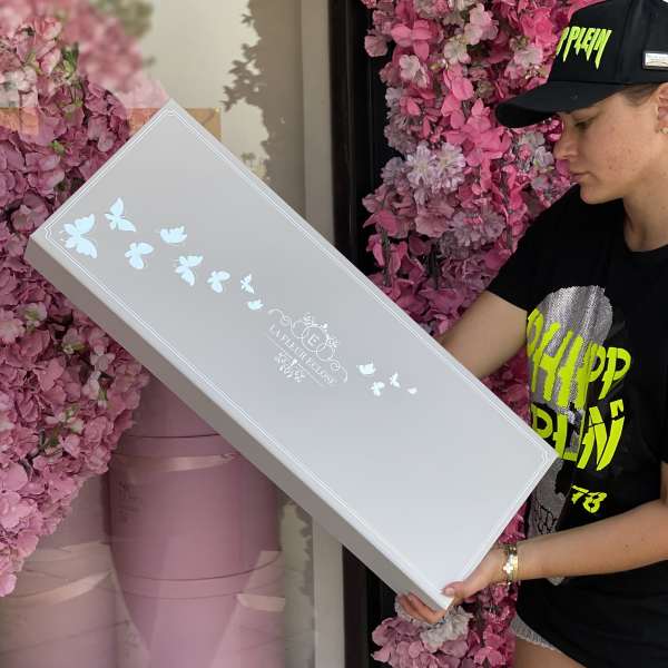 Woman holding a long white gift box in front of pink flower walls