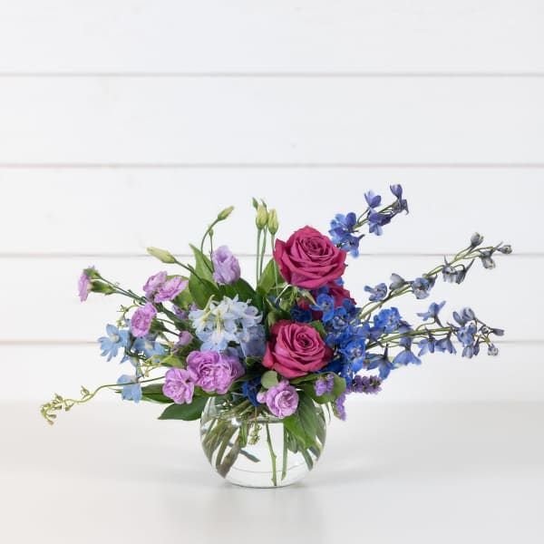 Mixed bouquet of pink roses, blue delphinium, and purple blooms in a glass vase