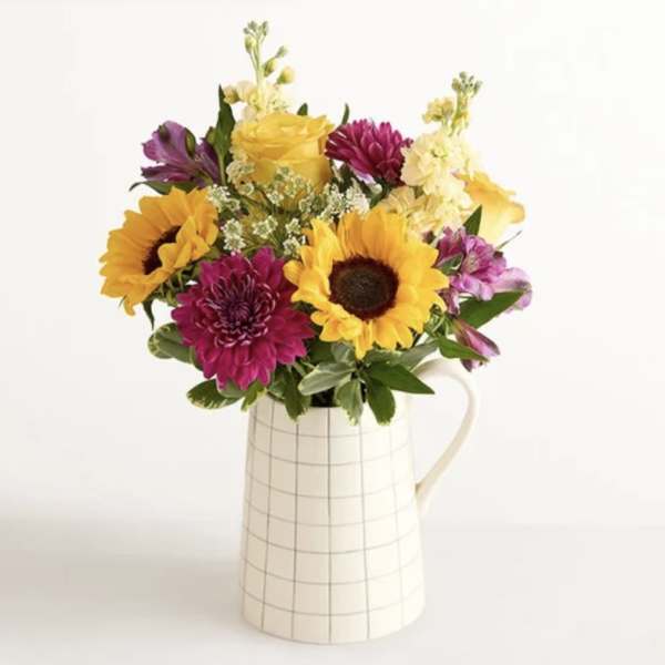 Bouquet of sunflowers, roses, and purple blooms in a white pitcher