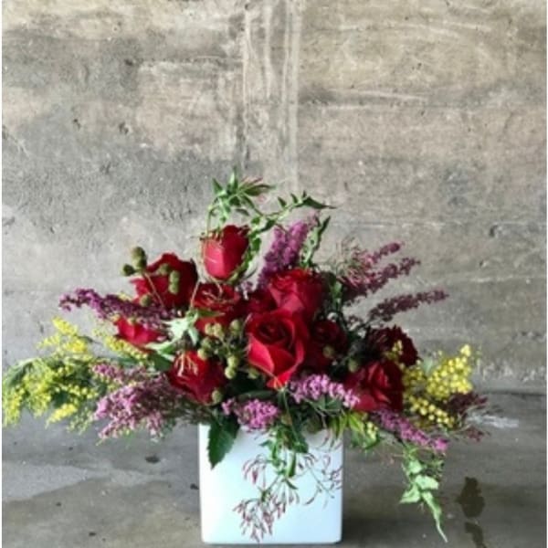 Red rose arrangement in a white square vase with purple and yellow filler flowers