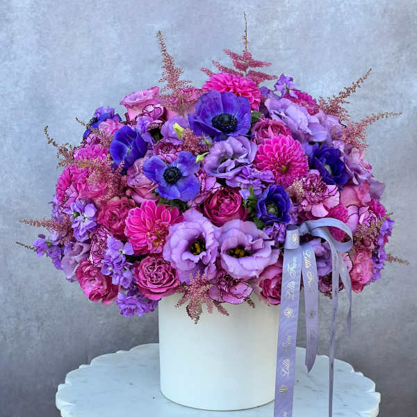 Purple and pink floral arrangement in a white box with ribbon tails