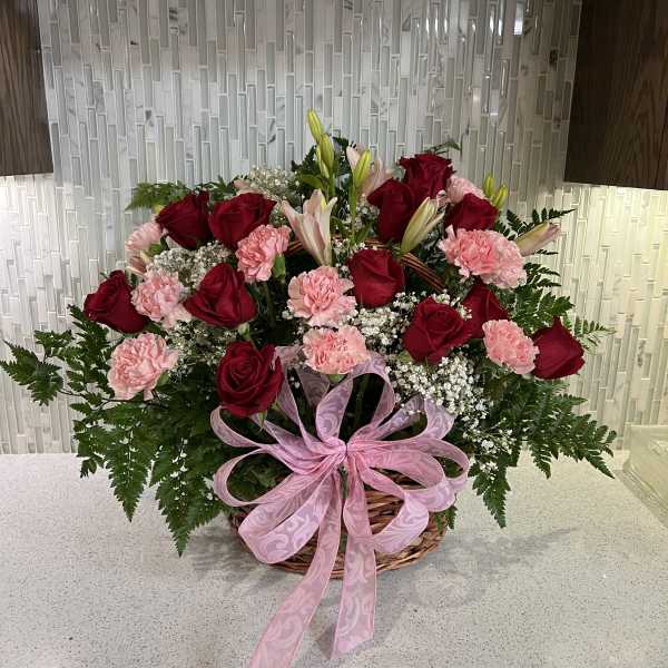 Basket arrangement of red roses, pink carnations, and lilies with a pink ribbon