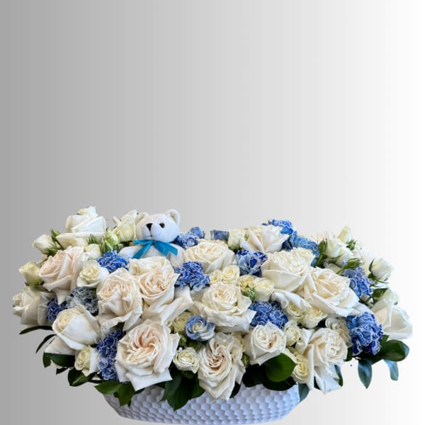 White rose arrangement with blue carnations and a teddy bear in a white basket
