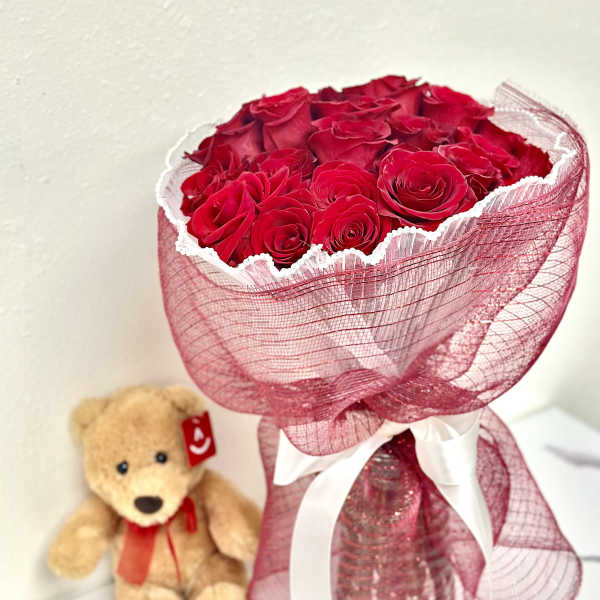 Bouquet of red roses wrapped in pink mesh with a teddy bear beside it