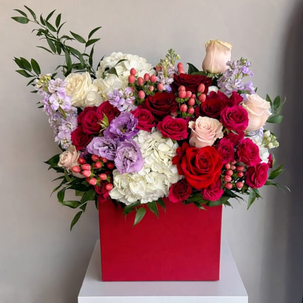 Lush arrangement of red and pink roses with white hydrangeas and purple blooms in a red square box