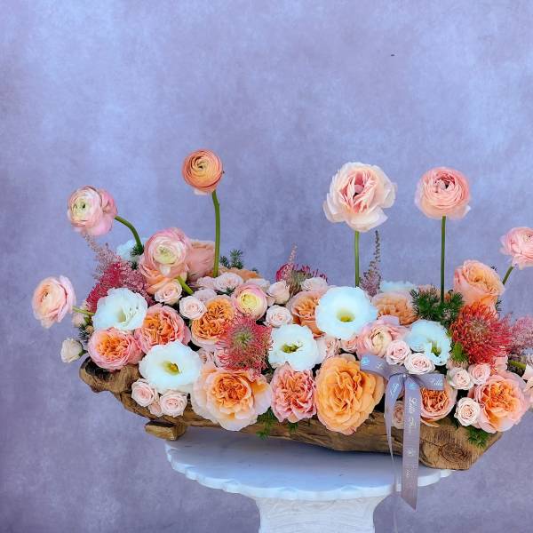 Low floral arrangement of peach, pink, and white blooms in a wooden container