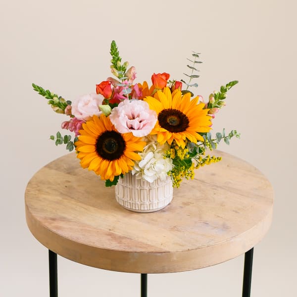 Small vase arrangement of yellow sunflowers, pink lisianthus and snapdragons in a white ceramic pot.