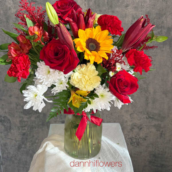 Mixed bouquet of red roses, lilies, sunflowers, and white daisies in a glass vase