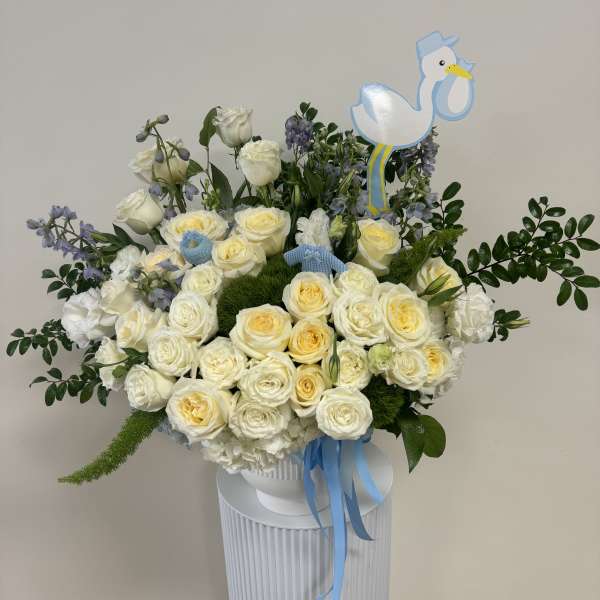 Large arrangement of cream roses and white flowers with blue ribbons and a stork pick in a white vase.