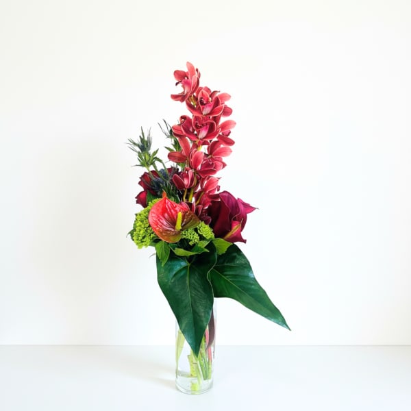 Tall modern arrangement of red orchids, calla lilies, and anthurium with large glossy green leaves in a clear glass vase
