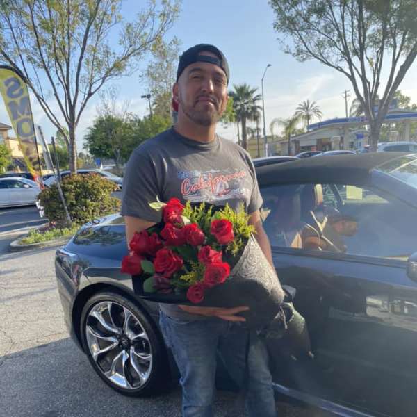Man holding a wrapped bouquet of red roses beside a dark car in a parking lot