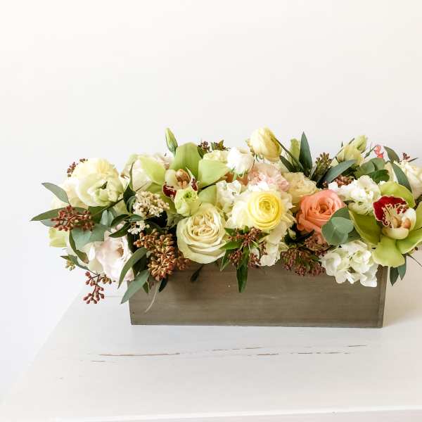 Low floral arrangement in a rectangular wooden box with cream, peach, and green blooms