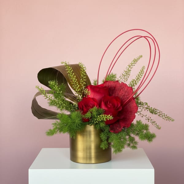 Red roses in a gold vase with green foliage and curved pink accents