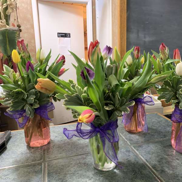 Four arrangements of mixed-color tulips in small glass vases with purple ribbon bows.