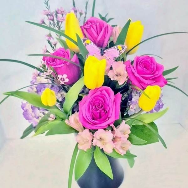 Arrangement of bright pink roses, yellow tulips, and pastel blooms in a black vase with a pink butterfly decoration