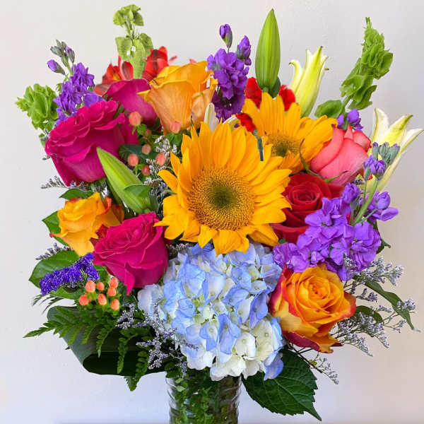 Tall mixed bouquet with sunflowers, roses, lilies, hydrangea, and purple stock in a clear glass vase.
