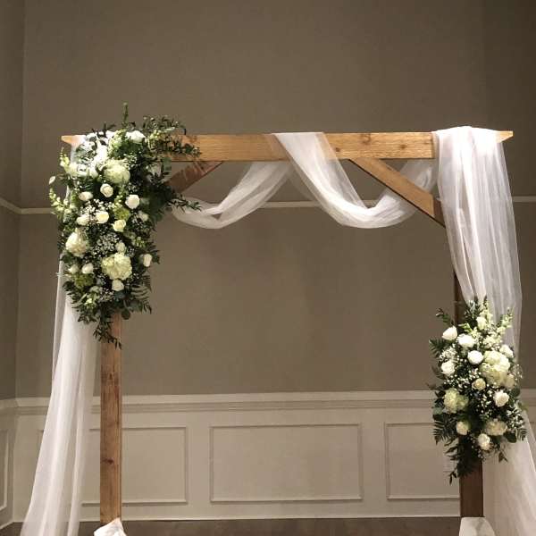 White floral arrangements on a wooden wedding arch with draped white fabric