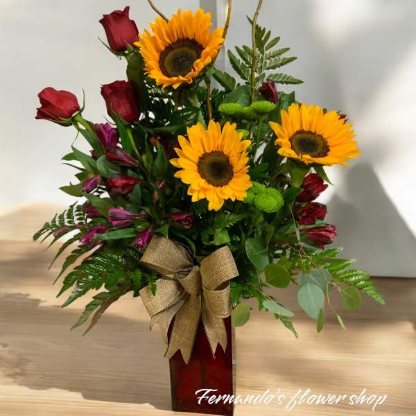 Arrangement of yellow sunflowers, red roses, and purple blooms in a red vase with a burlap bow