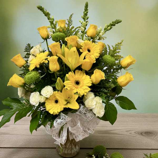 Yellow roses, lilies, and gerbera daisies in a glass vase with a white ribbon.