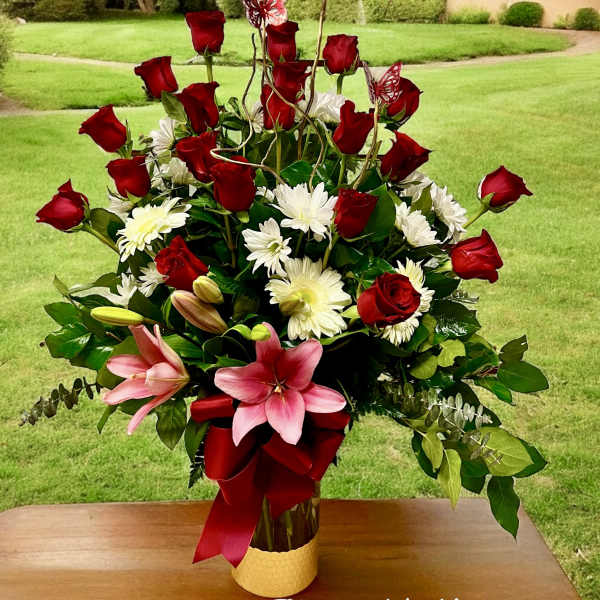 Tall arrangement of red roses, white daisies, and pink lilies in a glass vase with red bow
