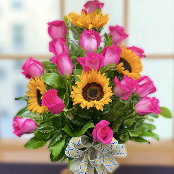 Arrangement of bright pink roses and yellow sunflowers in a glass vase with a patterned ribbon bow