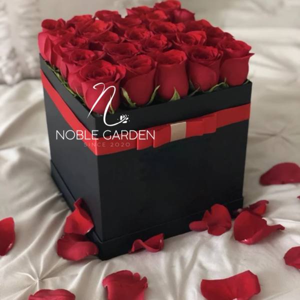 Red roses arranged in a black square gift box with rose petals around it