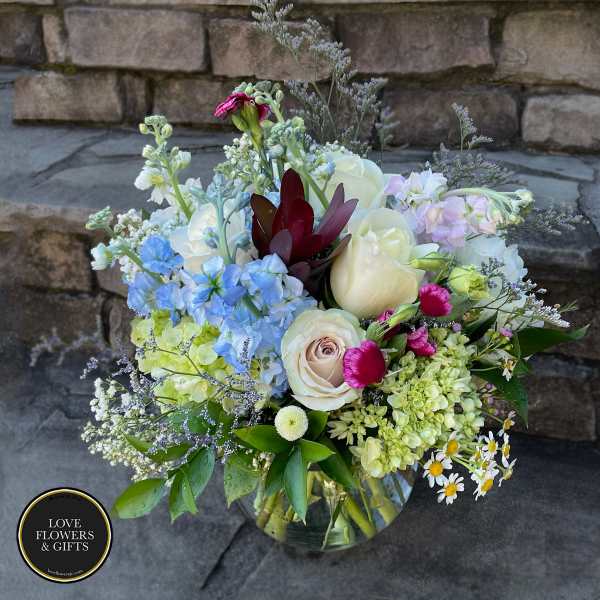 Mixed bouquet of cream roses, blue delphinium, green hydrangea and small daisies in a clear glass vase.
