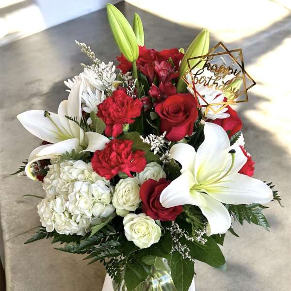 Red and white bouquet with lilies, roses, and a birthday topper in a glass vase