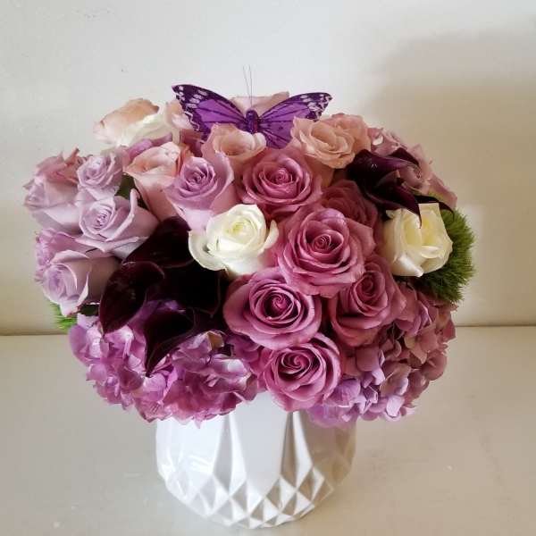 Pink and lavender roses arranged in a white vase with a purple butterfly topper