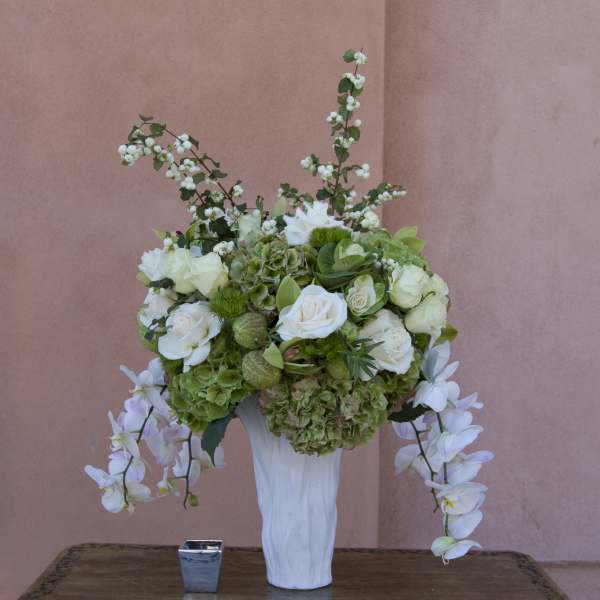 White and green floral arrangement in a tall white vase with trailing orchids
