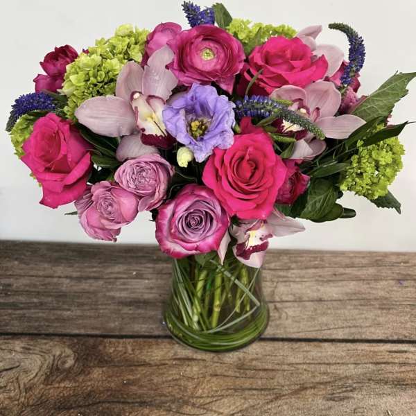 Bright pink and purple bouquet with roses, ranunculus, orchids, and hydrangea in a clear glass vase