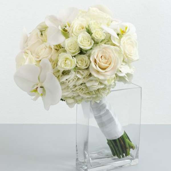 White bridal bouquet of roses, hydrangea, and orchids with ribbon-wrapped stems in a clear glass vase