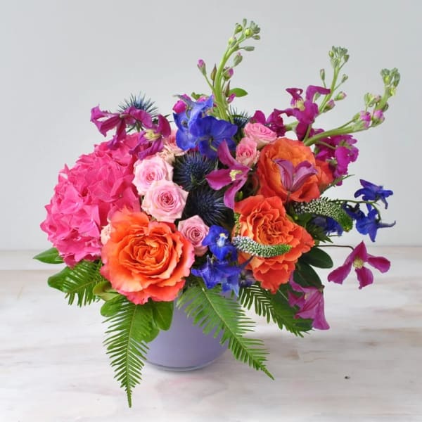 Bright mixed bouquet in a lavender vase with pink, orange, and blue flowers