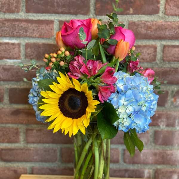 Bouquet of sunflowers, pink roses, blue hydrangeas, and orange tulips in a glass vase