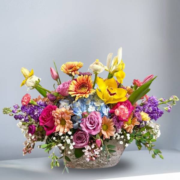 Colorful arrangement of roses, gerbera daisies, orchids, and mixed blooms in a low ceramic container