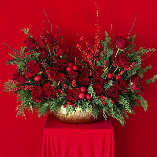 Red roses arranged in a gold vase with evergreen accents and red berries