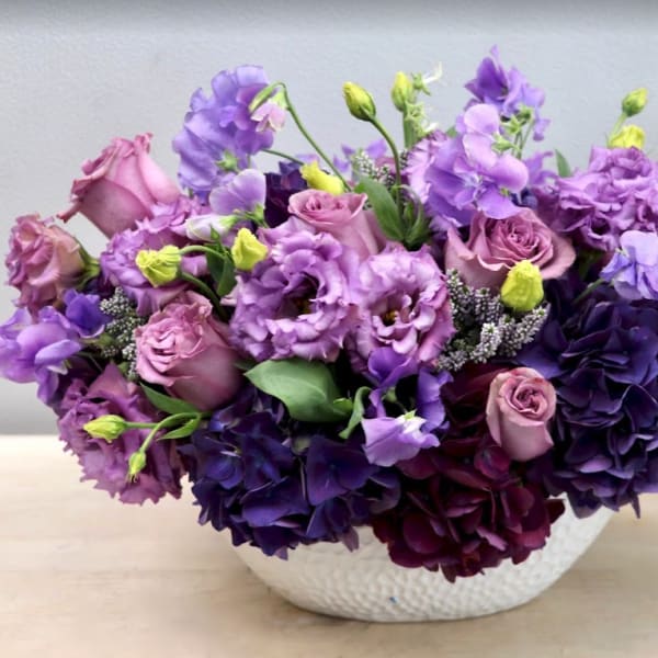 Low arrangement of purple and lavender roses and lisianthus in a white bowl vase